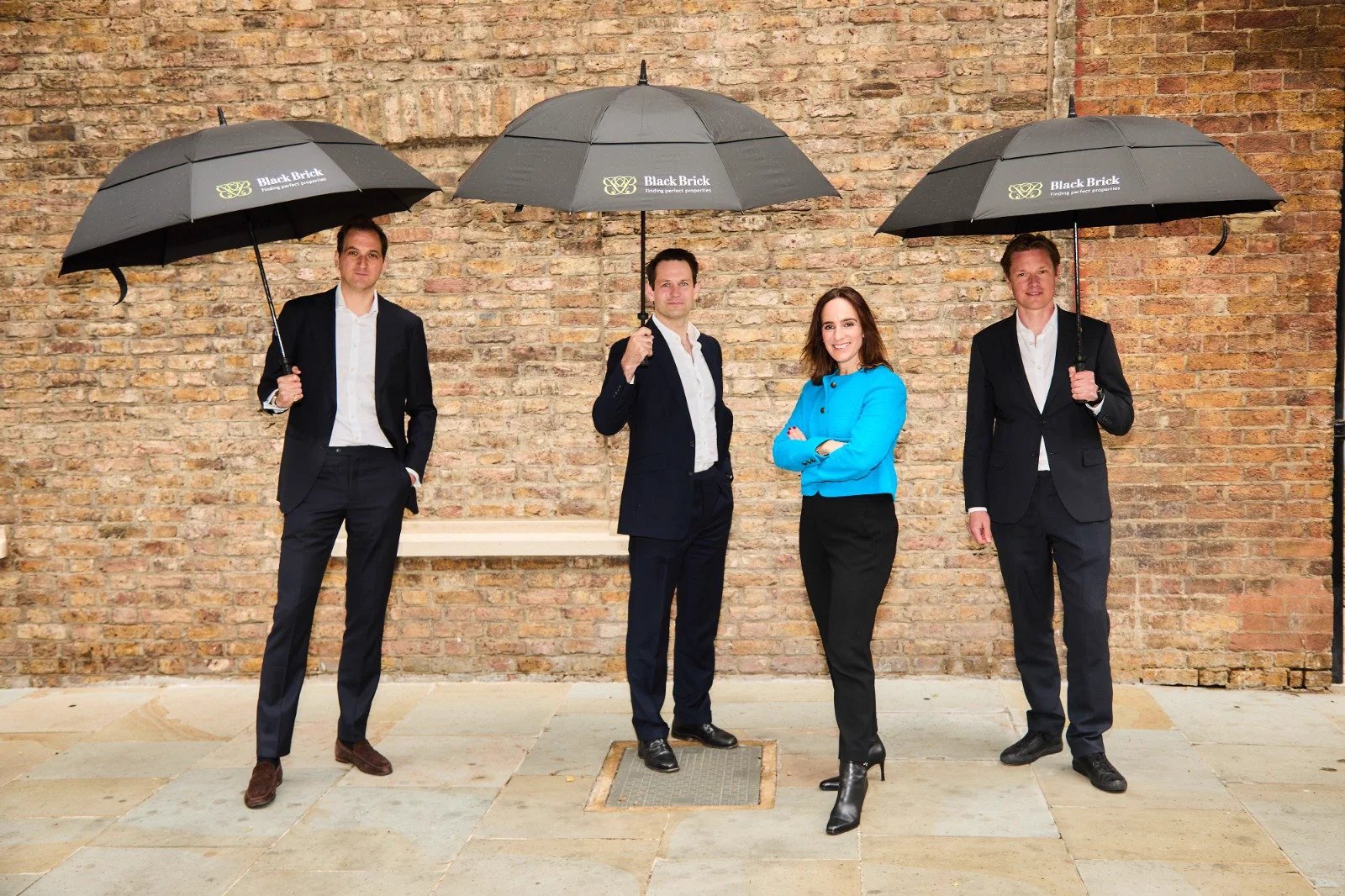 Four of the Black Brick team under branded umbrellas in front of a brick wall