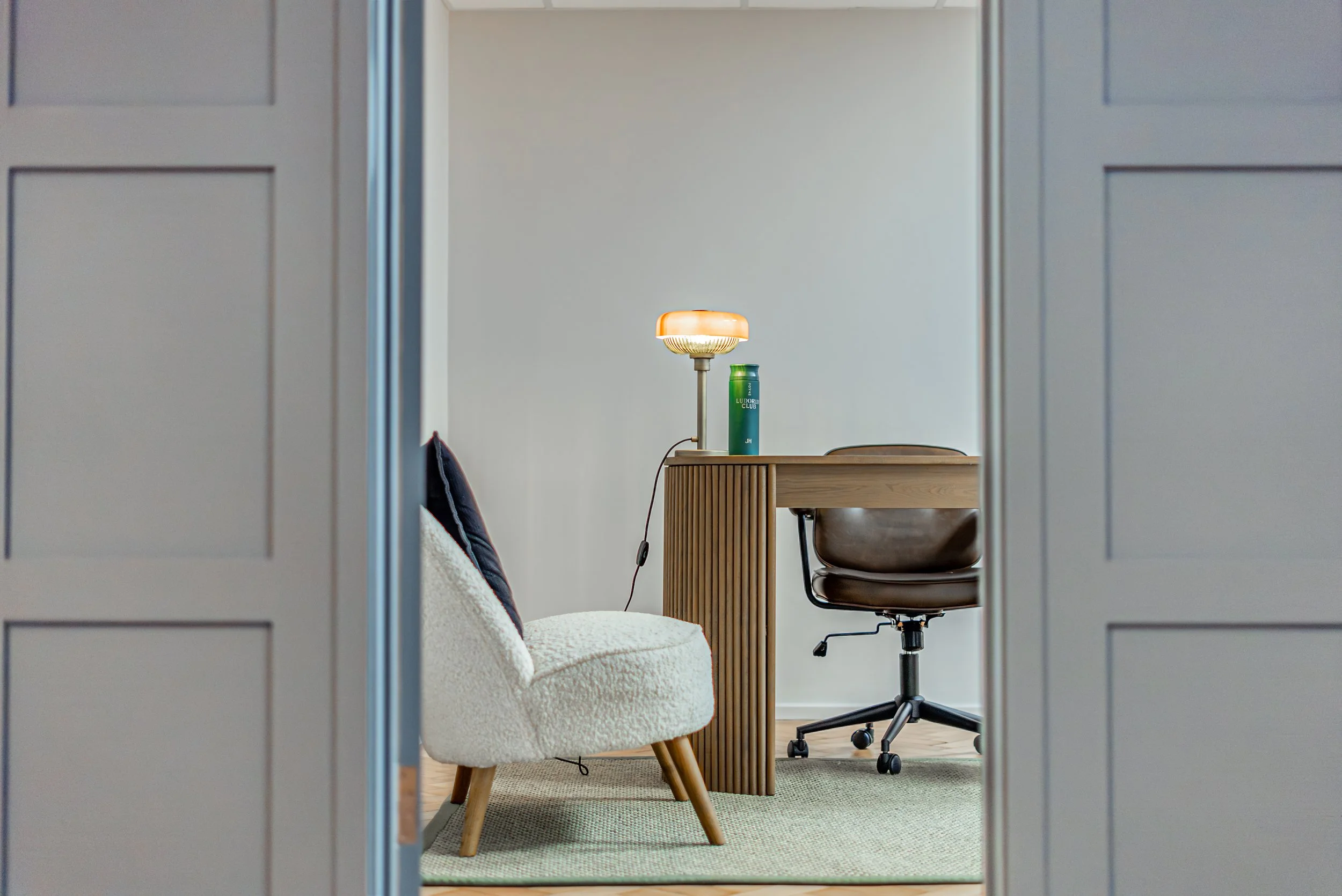 Looking through a half open panelled door into the consultation room: a boucle chair, a fluted desk and an amber lamp