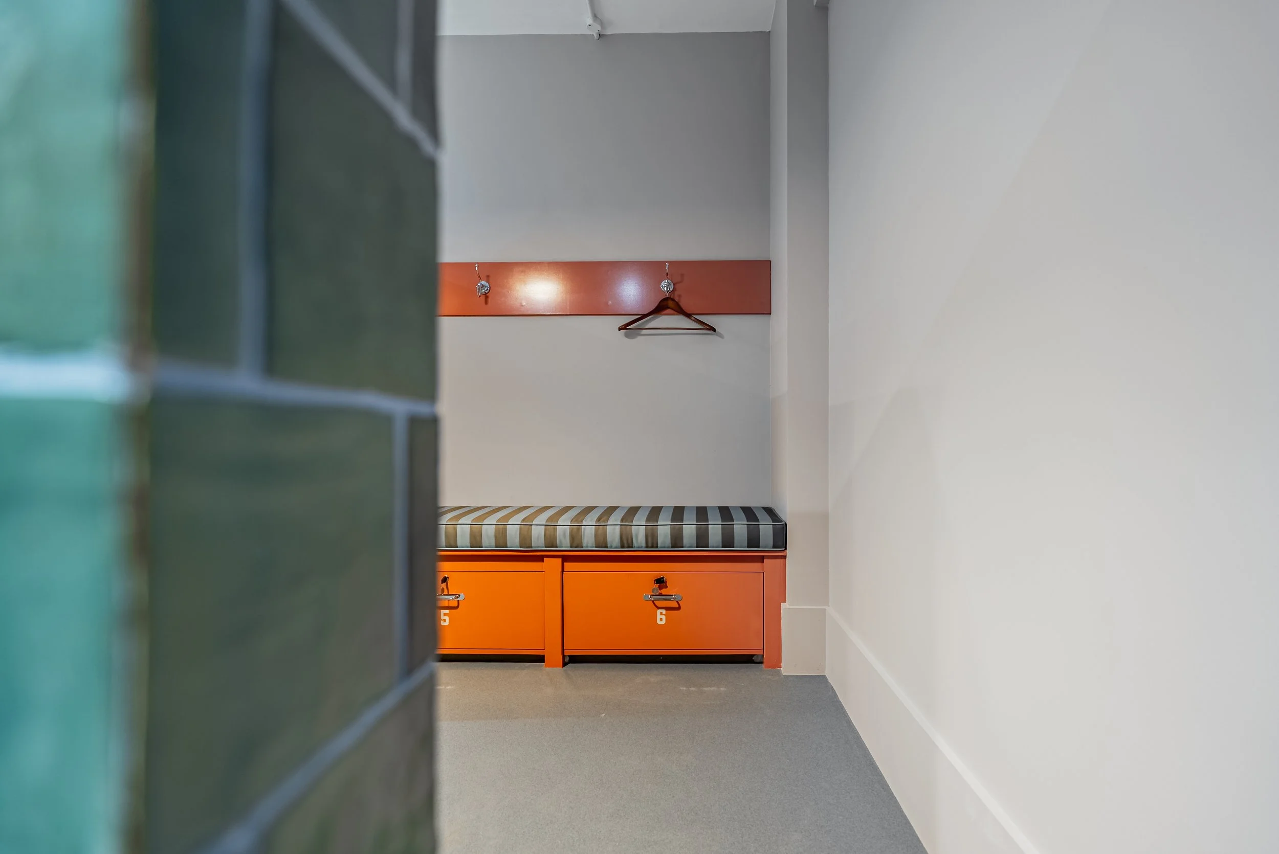 A glimpse past green glazed tiles into the locker room: orange drawers numbered five and six under a striped bench