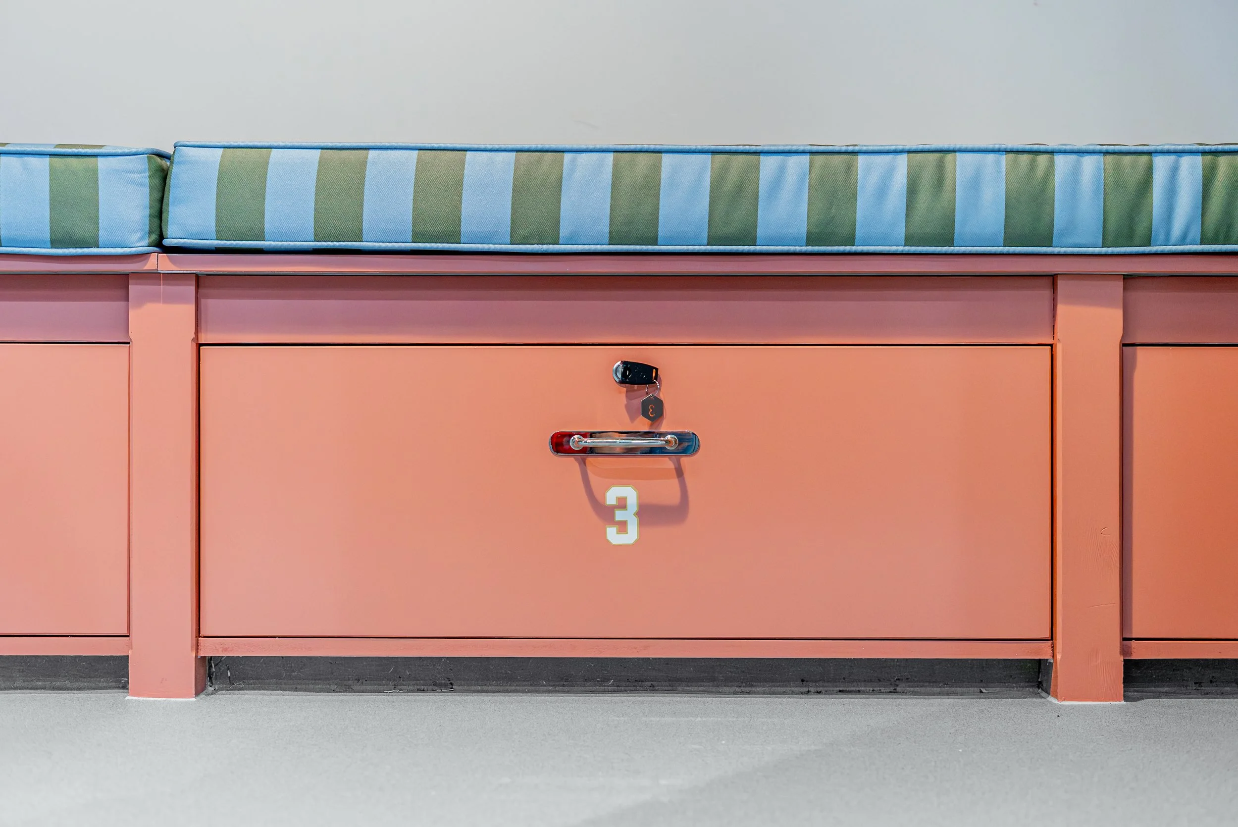 A coral painted locker drawer numbered three, under a blue and green striped bench cushion